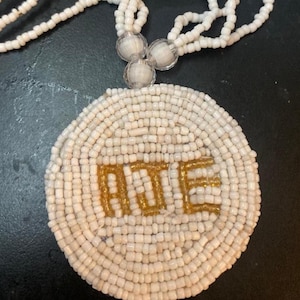 Puede incluir: Un collar de cuentas blancas con un colgante circular. El colgante presenta las letras doradas "ADE" en el centro. El collar tiene múltiples hilos de cuentas blancas y acentos redondos y transparentes.