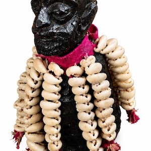 May include: A dark brown, carved wooden bust of a human head with intricate patterns, adorned with a bright pink fabric collar and multiple long, hanging strands of off-white cowrie shells that terminate in small, frayed red fabric tassels.