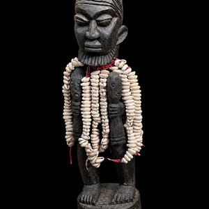 May include: A dark brown carved wooden figure standing on a circular base with etched wave patterns. The figure features a textured hairstyle, a beard, and is adorned with multiple strands of white cowrie shells draped over the shoulders.