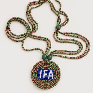 Puede incluir: Un collar de cuentas con un colgante circular. El colgante presenta las letras "IFA" en blanco sobre un fondo azul, rodeadas de cuentas verdes y marrones. Los hilos del collar son verdes y marrones.