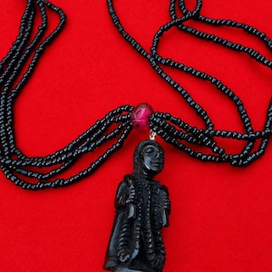Puede incluir: Un collar de múltiples hilos con pequeñas cuentas negras y cuentas de acento magenta translúcidas más grandes. Un colgante de figura negra tallada cuelga del centro, suspendido por un lazo de metal plateado unido a los hilos de cuentas.