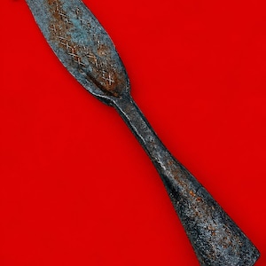 Puede incluir: Una herramienta de metal gris oscuro desgastada con una hoja ancha, plana y curva que presenta un patrón de rayas cruzadas y un mango cónico. La superficie muestra una oxidación y óxido significativos, con un acabado texturizado e irregular.
