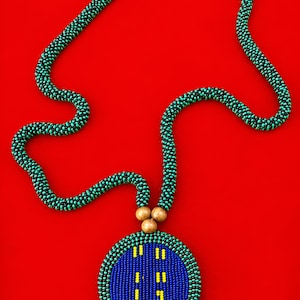 Puede incluir: Un collar largo con una cuerda tubular hecha de pequeñas cuentas negras y verde azulado. El colgante es un disco circular grande y plano con un centro azul, detalles verticales amarillos y un borde de cuentas negras y verde azulado.