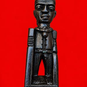 Peut inclure: Une figurine en bois sculpté brun foncé représentant un personnage debout portant un chapeau structuré et un vêtement à col avec des détails verticaux. La figurine présente des traits du visage marqués, dont de grands yeux et un nez défini, sur un fond rouge uni.