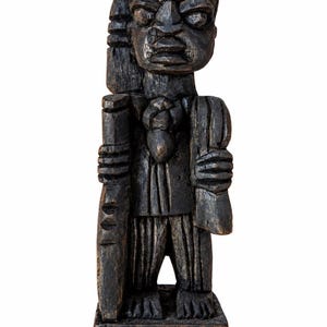May include: A dark brown, hand-carved wooden figurine standing on a rectangular base. The figure features a stylised head with prominent facial features, a textured torso, and vertical lines on the lower garment, holding a long, narrow object.