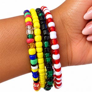 May include: A set of four elastic bracelets made of small, round plastic beads. One bracelet features a multicoloured pattern, another is solid yellow, the third is black and green, and the fourth alternates between red and white beads.