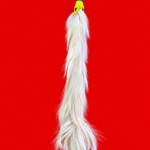 Puede incluir: Una cola de animal larga de color blanco roto con un mango de cuentas que presenta un patrón geométrico amarillo y verde. El mango está rematado con un lazo trenzado amarillo y verde para colgar, creando un accesorio decorativo texturizado.