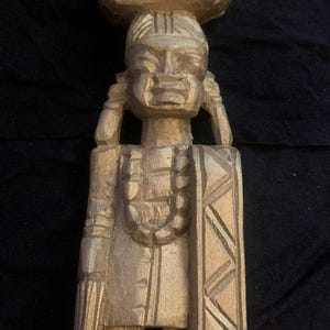 Puede incluir: Estatua de madera tallada de una persona que lleva un objeto grande sobre la cabeza. La figura tiene un rostro detallado, un collar y una prenda estampada. La estatua es de color marrón claro y se apoya sobre una base rectangular.