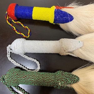 May include: Three beaded ceremonial whisks. The handles are cylindrical and covered in colorful beads. One is red, blue, and yellow, another is white, and the third is green and brown. Each has a white, fluffy tail.
