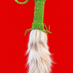 Könnte beinhalten: Ein dekoratives Ornament mit einem zylindrischen Griff, der mit kleinen grünen und gelben Perlen bedeckt ist und von einer passenden Perlenschlaufe gekrönt wird. Ein langes, fließendes Büschel aus hellbeigem und weißem Kunstfell ist an der Basis befestigt.