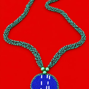 Puede incluir: Un collar largo con una cadena tubular de cuentas que alterna colores turquesa y marrón oscuro. La cadena se conecta a un gran colgante circular hecho de cuentas azules con detalles verticales en blanco y tres cuentas verdes en la parte superior.