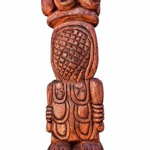 Puede incluir: Una escultura de madera tallada a mano que presenta una figura central con una cabeza ovalada de patrón de rejilla texturizada, rematada por dos rostros más pequeños. El torso muestra tallas en forma de U y bandas horizontales.
