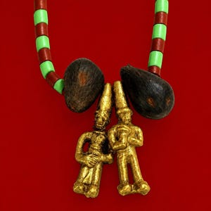 May include: A necklace featuring two gold-colored metal human-shaped pendants suspended between two dark brown, textured, teardrop-shaped beads. The necklace strand consists of alternating light green and dark red cylindrical beads.