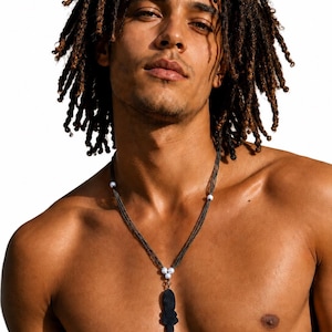 May include: A multi-strand dark grey necklace featuring three small white spherical beads arranged in a triangular cluster at the centre, connected to a singular, elongated, matte black pendant with a distinct, symmetrical, notched silhouette.