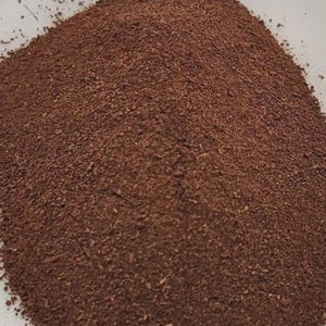 Puede incluir: Primer plano de un montón de material finamente molido, de color marrón oscuro. La textura parece granular y uniforme, lo que sugiere un tamaño de partícula constante. El color es un marrón rico y terroso, sin variaciones visibles.