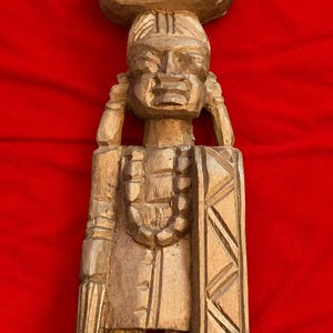 Puede incluir: Una escultura de madera tallada a mano de color marrón claro que representa una figura que lleva un recipiente grande y redondeado sobre la cabeza. La figura presenta un collar de cuentas, cabello texturizado y una prenda rectangular.