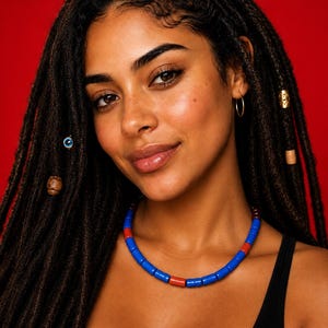 Puede incluir: Un collar con un patrón repetitivo de cuentas cilíndricas azules y rojas. La joya se lleva con rastas de color marrón oscuro adornadas con pequeñas cuentas de metal y madera, incluyendo un amuleto de ojo turco azul y detalles dorados.