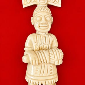 Pode incluir: Um pingente esculpido em tom off-white apresentando uma figura em pé com um grande adorno de cabeça, olhos arregalados e uma túnica texturizada. A figura segura um objeto cilíndrico à frente do corpo. Um passador de metal prateado está fixado no topo.