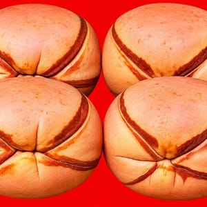 May include: Four round, light tan baked bread rolls arranged in a two-by-two grid against a solid red background. Each roll features a distinct, dark brown cross-shaped indentation scored into the top surface, creating a textured appearance.