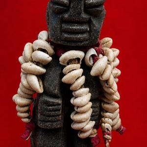 Pode incluir: Uma estatueta de pedra cinza-escura com cabeça arredondada e traços faciais esculpidos. A figura é adornada com vários fios de conchas de búzios em tom off-white, enfiados em cordões de tecido vermelho que pendem ao redor do pescoço.