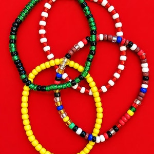Puede incluir: Tres pulseras circulares hechas con pequeñas cuentas redondas de plástico. Una pulsera es de color amarillo sólido, otra alterna rojo, blanco y negro, y la tercera presenta una mezcla de cuentas verdes, negras, rojas, blancas, azules y transparentes.