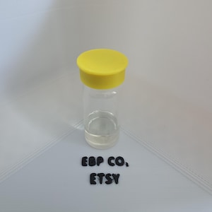 Peut inclure: Un flacon en verre transparent avec un bouchon jaune. Le flacon contient un liquide clair. Le texte "EBP CO., ETSY" est en dessous du flacon. Le fond est blanc.