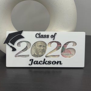 May include: A white rectangular sign featuring the black text Class of and the name Jackson. The numbers 2026 are cut out, revealing US currency behind them. A black graduation cap icon is attached to the left side of the white sign.
