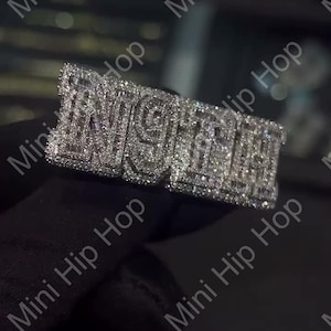 May include: A silver-toned ring with the word "NIGHT" spelled out in block letters, encrusted with small, sparkling stones. The ring is set against a dark background, highlighting its reflective surface. The ring is a piece of jewelry.