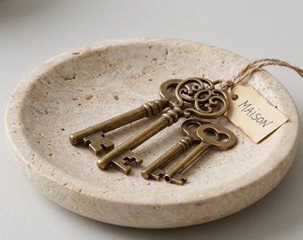 以下が含まれることがあります： ベージュ色の多孔質な石で作られた丸いトレイに、アンティーク調の真鍮製の鍵が3本置かれています。鍵には麻紐で小さな長方形の薄茶色の紙タグが結び付けられており、表面には黒いインクで「MAISON」という文字が書かれています。