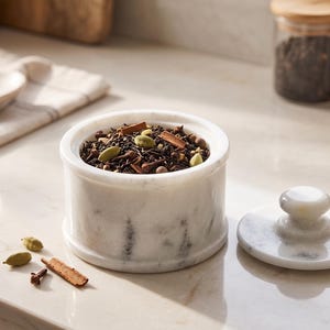 May include: A white marble canister with a matching lid, filled with a blend of loose leaf tea, green cardamom pods, cinnamon sticks, and cloves. A few loose cardamom pods, a cinnamon stick, and a clove rest on the surface next to the canister.