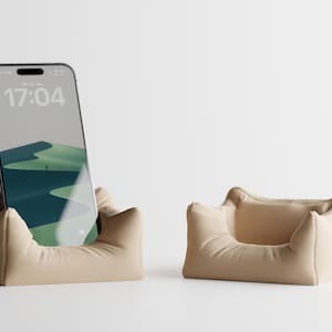 May include: Two beige phone stands. One holds a smartphone displaying a landscape image and the time 17:04. The other stand is empty. Both stands have a soft, pillow-like appearance.
