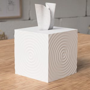 May include: A white square tissue box with a concentric circle design on the sides. Two white tissues are emerging from the top. The box is sitting on a wooden surface.