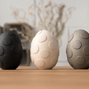 May include: Three decorative egg-shaped objects with a textured surface and large, raised circular patterns. The eggs are in black, off-white, and taupe colors, and are arranged on a wooden surface. The background is blurred, suggesting a studio or home setting.