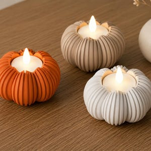 May include: Three pumpkin-shaped candle holders in orange, beige, and white. Each holder features a ribbed exterior and contains a white LED tea light with a glowing flame-shaped bulb, creating a warm light effect for seasonal decor.