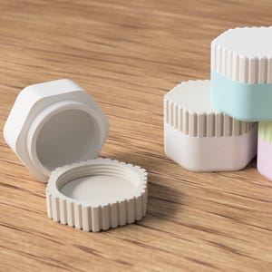 May include: White, hexagonal cosmetic jars with ribbed lids. One jar is open, revealing the interior. Other jars have pastel-colored bands in blue, green, and pink. The jars are on a wooden surface.