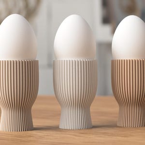 May include: Three white eggs sit atop wooden egg cups with vertical grooved detailing. The egg cups are in shades of beige and brown, arranged on a light-colored wooden surface. The background is blurred.
