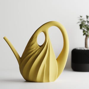 May include: A yellow watering can with a unique, sculptural design. The can features a large handle, a long spout, and a decorative, swirling pattern on the body. A small potted plant is in the background.