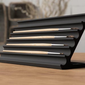 May include: A black, tiered brush holder with five paint brushes. The holder has a curved design and is made of a matte black material. The paint brushes have light-colored wooden handles and dark bristles.