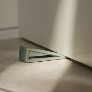 May include: A light green door stop in a wedge shape, positioned on a light-colored floor. The door stop is made of a solid material and is designed to prevent a door from closing. The wedge shape has a textured top.