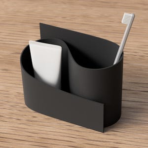 May include: A black toothbrush holder with a curved design, holding a white tube of toothpaste and a white toothbrush. The holder is made of a matte material and sits on a wooden surface.