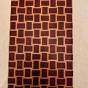 May include: Rectangular wooden chopping board with a mosaic pattern. The board has a repeating design of dark reddish-brown and light brown rectangular blocks. The wood grain is visible, adding texture to the surface.