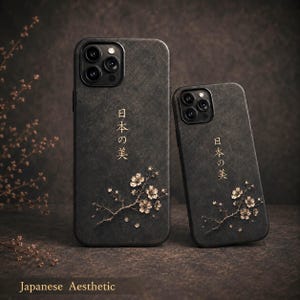 Könnte beinhalten: Zwei dunkelgraue Smartphone-Hüllen mit strukturierter Oberfläche, goldenen Kirschblütenzweig-Illustrationen und dem vertikal aufgedruckten japanischen Text 日本の美. Jede Hülle hat einen Ausschnitt für die Dreifachkamera.