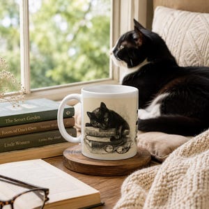 May include: A white ceramic mug featuring a black cat illustration resting on books, with eyeglasses. The mug sits on a wooden coaster, next to a stack of books and a black and white cat. A cozy scene with a window in the background.