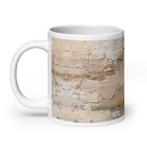 May include: White ceramic coffee mug featuring an abstract design with textured layers of beige, pink, and yellow. The mug has a white handle and a glossy finish. The design resembles a close-up of a textured wall.