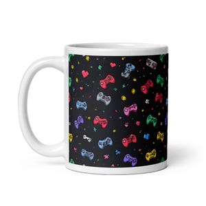 May include: White ceramic mug featuring a repeating pattern of colorful video game controllers, hearts, and stars on a black background. The controllers are in various colors, including red, blue, green, and purple.