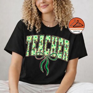 Puede incluir: Camiseta negra con la palabra "TEACHER" en un diseño verde y estampado de leopardo, adornada con tréboles. Debajo de la palabra hay un lazo verde y rosa. La camiseta es de cuello redondo.
