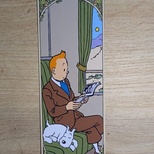 May include: A rectangular bookmark featuring a colorful illustration of a man with orange hair wearing a brown suit and blue tie, sitting on a green seat while reading a book. A small white dog is curled up asleep on the seat beside him.