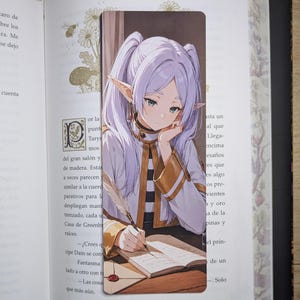 Puede incluir: Un marcapáginas rectangular con una ilustración estilo anime de un elfo de pelo violeta claro y orejas puntiagudas, que viste una chaqueta blanca y dorada con cuello, escribiendo en un libro con una pluma mientras apoya la mano en su barbilla.