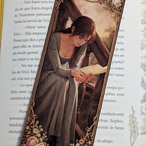 Puede incluir: Marcapáginas rectangular con una ilustración vintage de una mujer leyendo una carta. El marcapáginas tiene un marco dorado con detalles florales y de volutas. La mujer lleva un vestido largo gris.