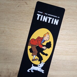May include: A rectangular black bookmark featuring the white text HERG&Eacute; THE ADVENTURES OF TINTIN at the top. Below the text, a yellow oval contains an illustration of Tintin in a red jacket and brown pants, with his black silhouette and Snowy the dog.